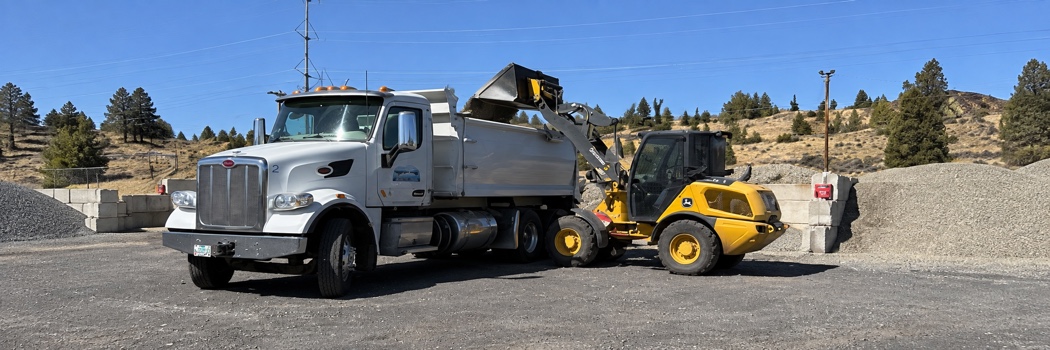 River’s Edge dump truck being loaded by a yellow skid-steer at the gravel yard